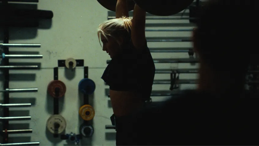 Silhouette of a woman lifting a barbell overhead in a gym with weight plates on racks in the background.
