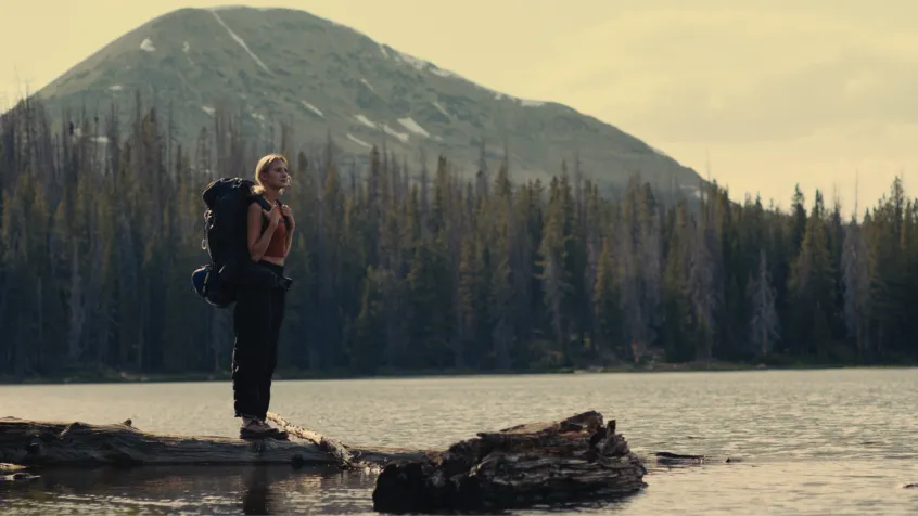 Hiker with a large backpack standing on a log by a lake with forest and mountain in the background at sunset.