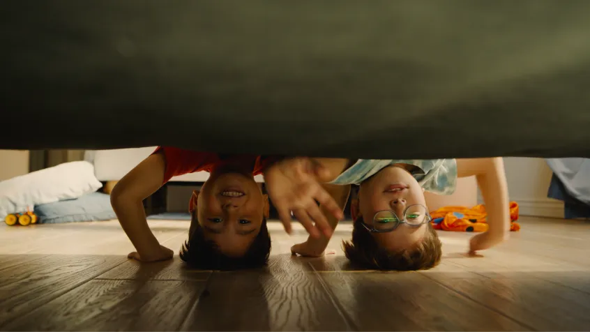 Two children lying on the wooden floor looking under a piece of furniture, one smiling and the other wearing glasses.