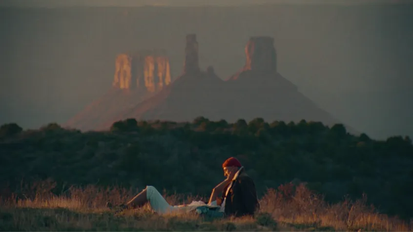 Person with a red headscarf sitting on the grass, looking at another person lying down, with large rock formations in the background at sunset.