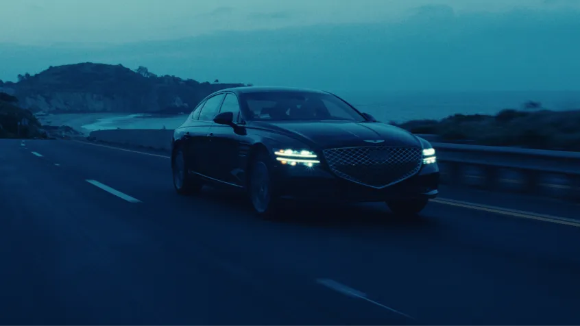 Black luxury sedan driving on a coastal road at dusk with headlights on.