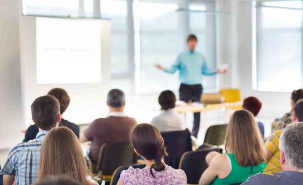 A group of people seated in rows attentively listening to a presenter in a bright room with large windows, where the presenter stands in front of a projection screen and gestures with arms.