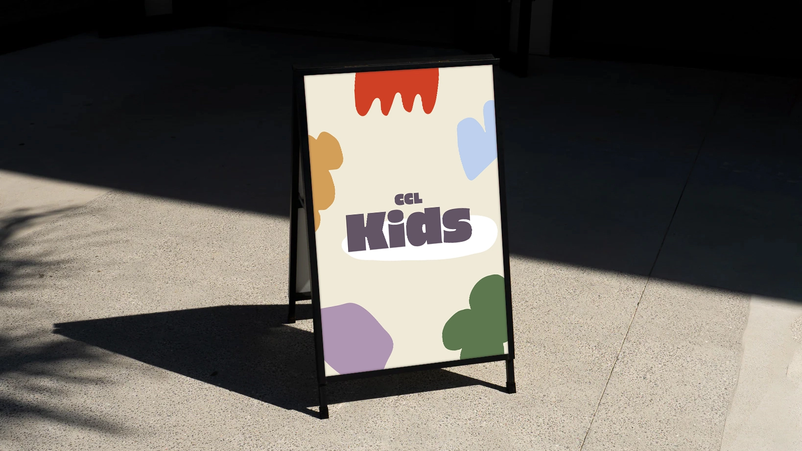 A-frame sign on pavement with colorful abstract shapes and text 'CCL Kids' in block letters.