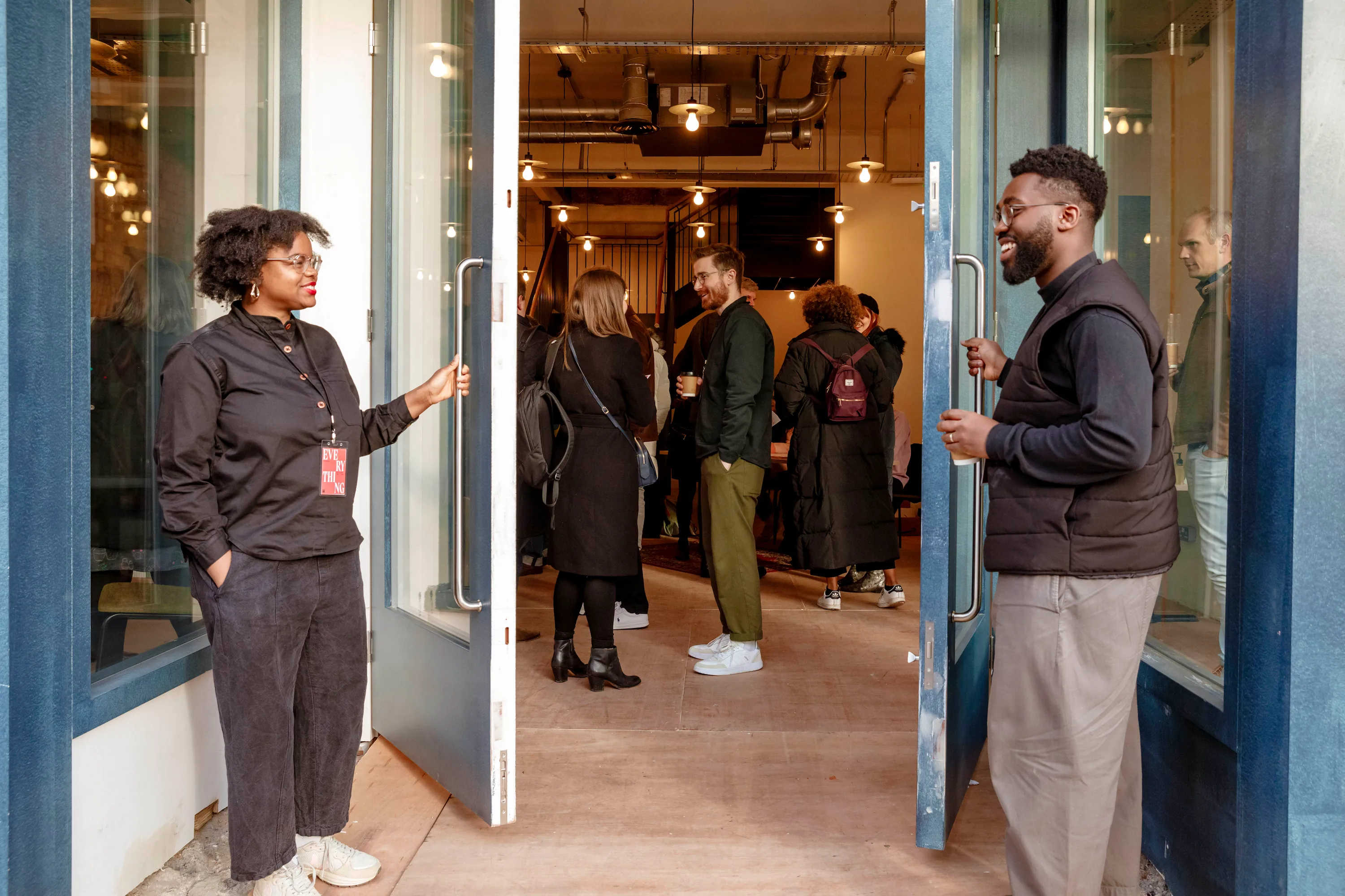 Two people hold open doors welcoming a crowd inside a warmly lit building event space.