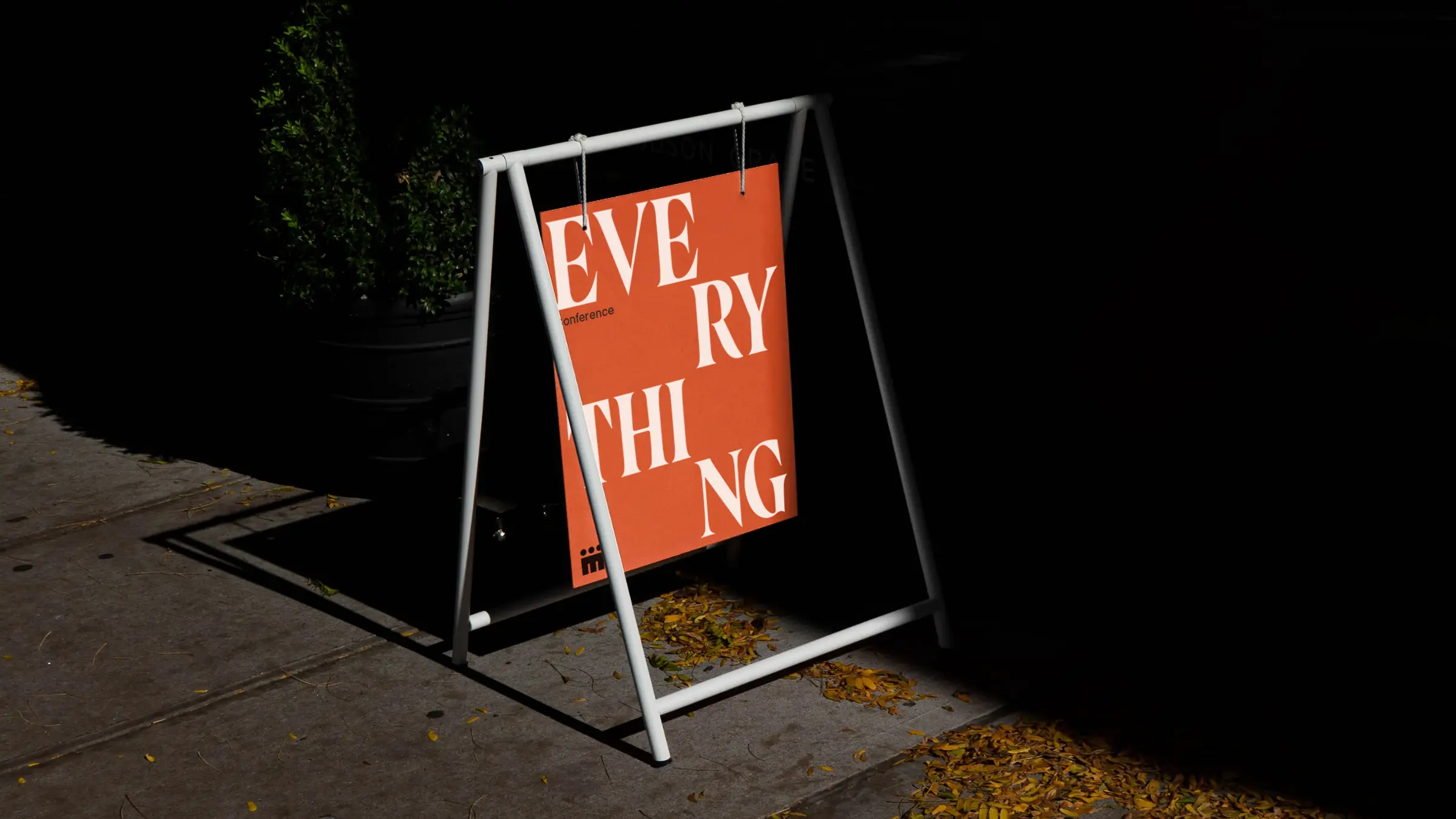 White metal frame holds an orange sign with scattered cream letters spelling 'EVERYTHING' on sidewalk.