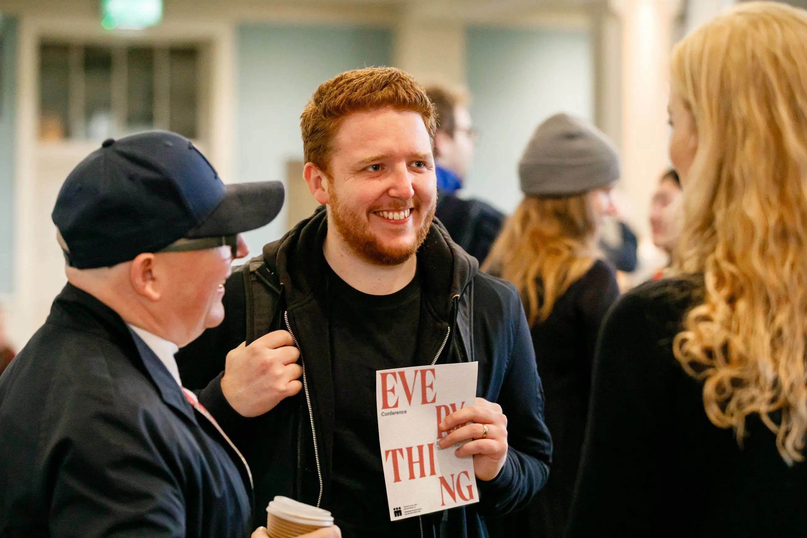 Smiling man holding a conference booklet talking with two people indoors at a social event.