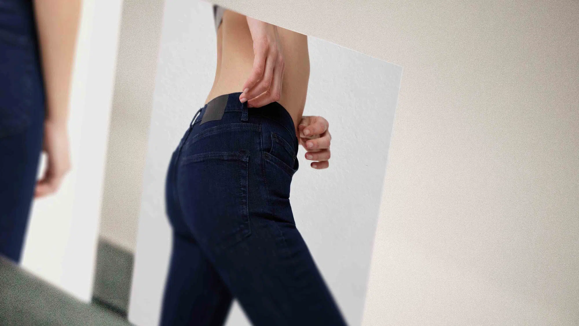 Person wearing dark blue jeans seen from the side adjusting the waistband in front of a mirror.