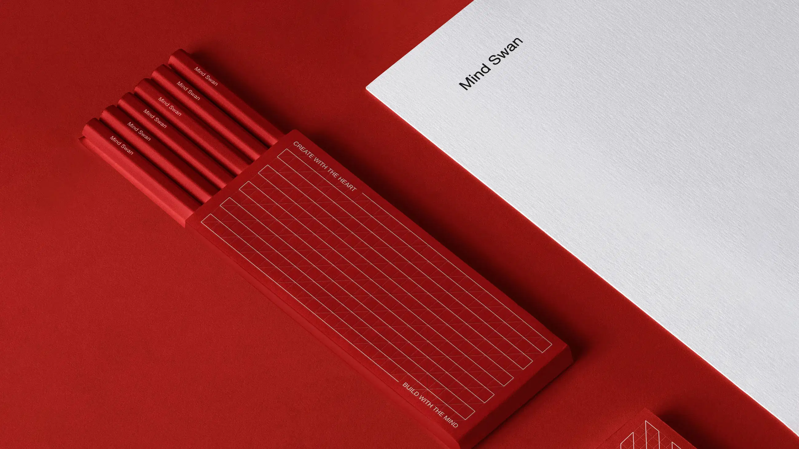 Set of six red pencils labeled 'Mind Swan' in a matching red box on a red surface next to white paper also labeled 'Mind Swan'.