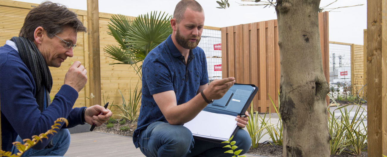 Two men outdoors examining plants, one holding a folder and the other holding a pen.