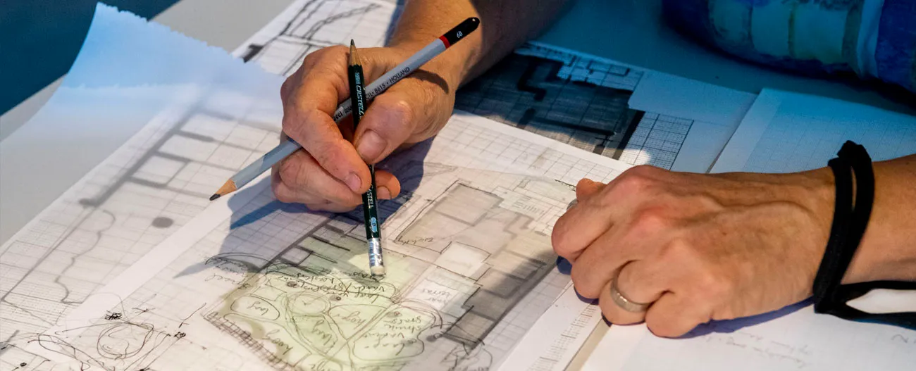 Person’s hands holding pencils and working on architectural or landscape design plans with notes and sketches on tracing paper.