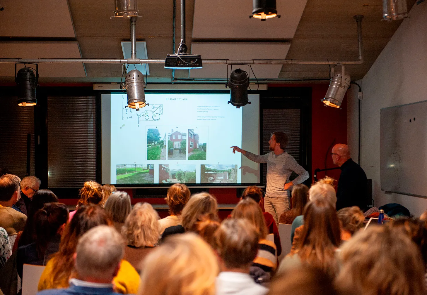 Man giving a presentation to an audience with images and text projected on a screen in a dimly lit room.