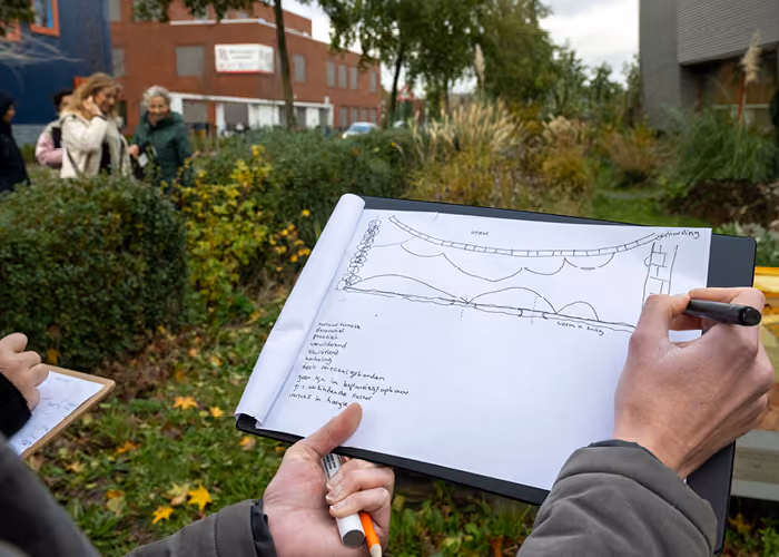 Person sketching a landscape plan on paper outdoors with greenery and buildings in the background.