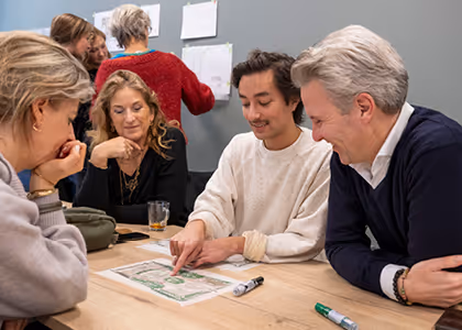 Five adults sitting and standing around a table, engaging in discussion over a document with markers on the table.