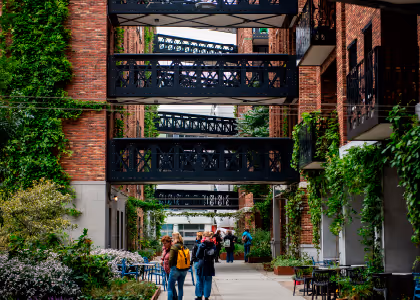 Pedestrian walkway between red brick buildings with multiple black iron balconies and greenery hanging from walls, with people walking.