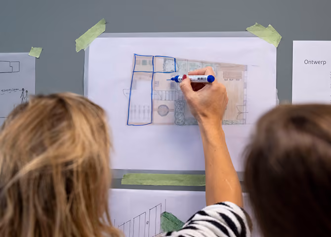 Person drawing blue outlines on a taped architectural floor plan while another person observes.