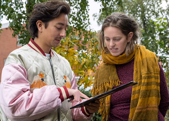 Two people outdoors looking at a clipboard, one pointing at the paper, surrounded by green and yellow foliage.
