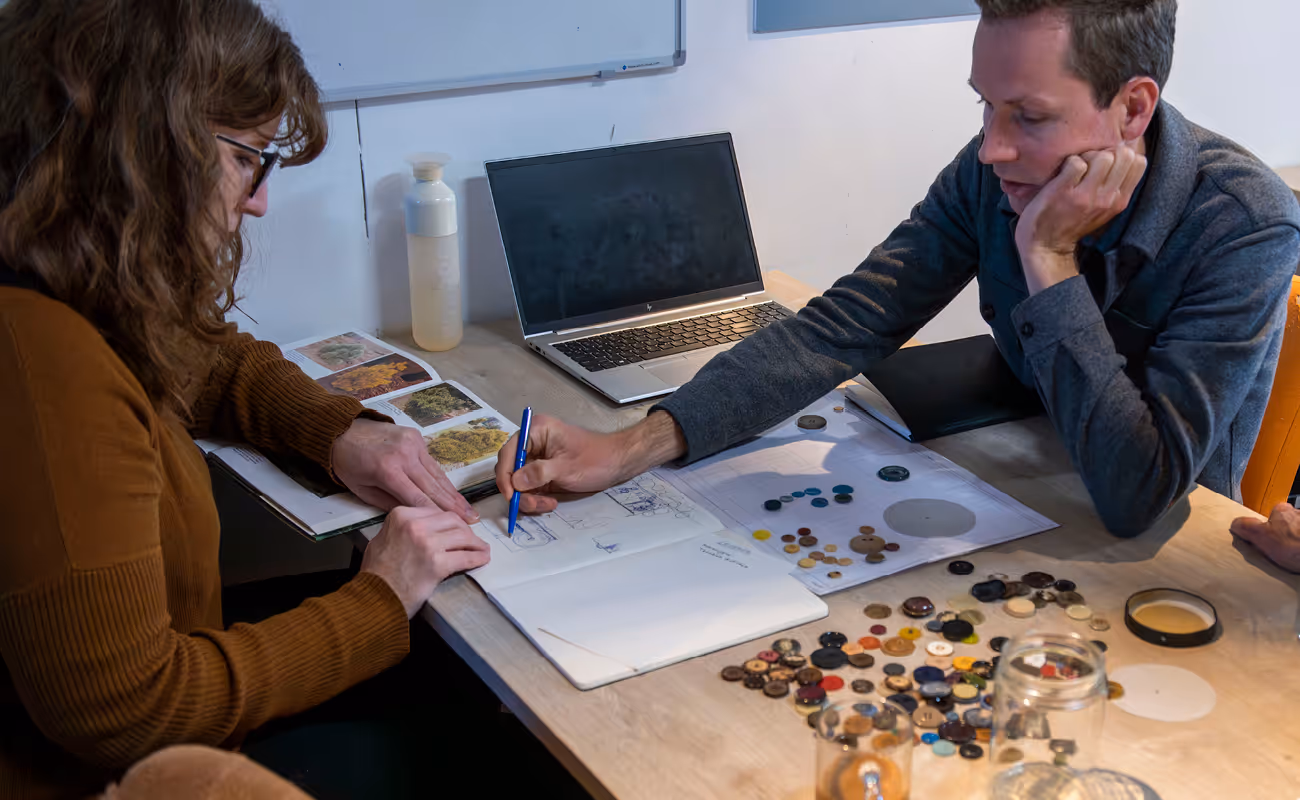 Two people sitting at a table with a laptop, sketching and examining a book with plant images, surrounded by various buttons and papers.