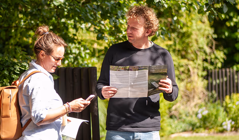 A man and a woman outdoors with maps, the woman using a smartphone and the man looking at the map, surrounded by greenery.