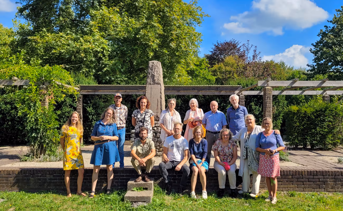 Group of fourteen people posing outdoors in front of a stone monument and greenery under a sunny blue sky.