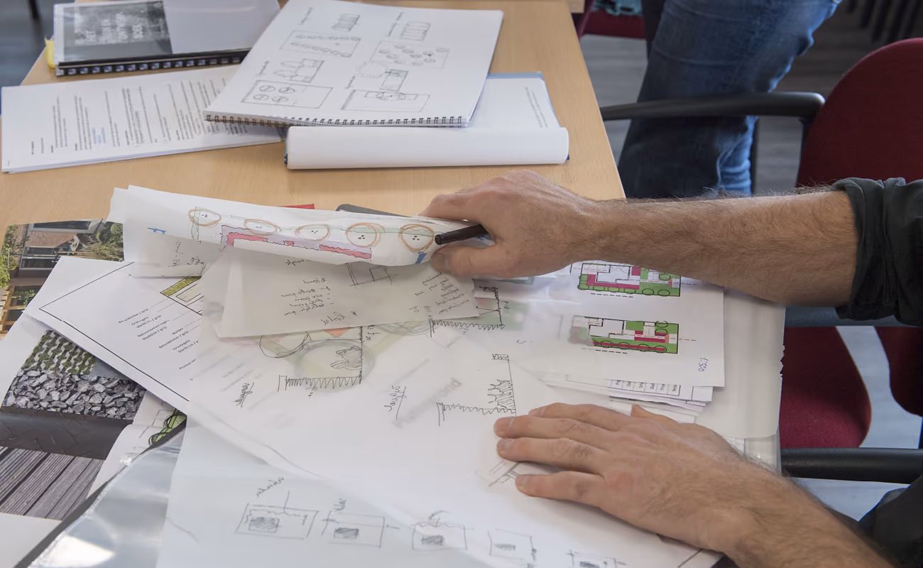 Person reviewing architectural and landscape design sketches and plans spread on a wooden table.