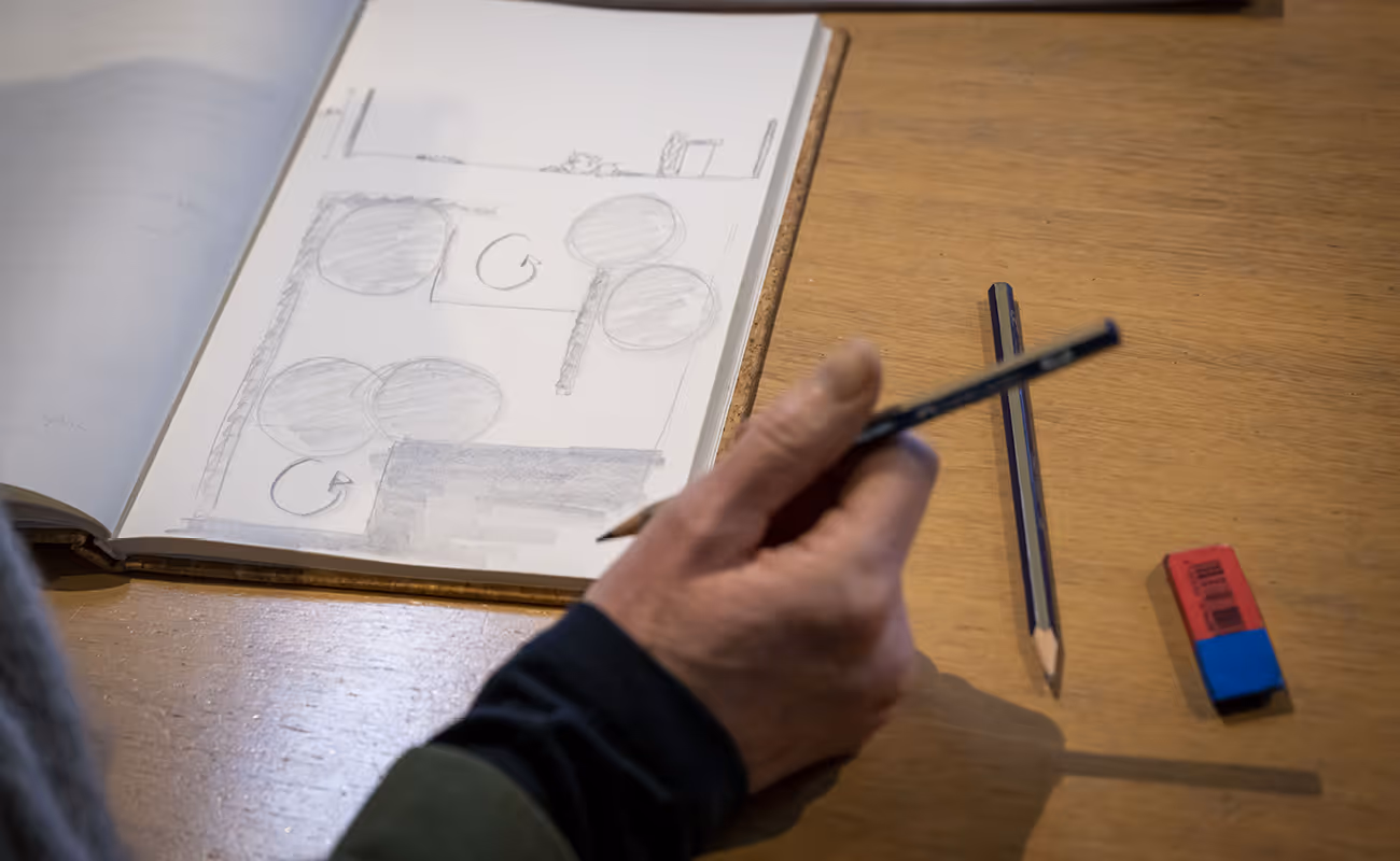 Hand holding a pencil above a sketchbook with geometric pencil drawings on a wooden table next to another pencil and eraser.