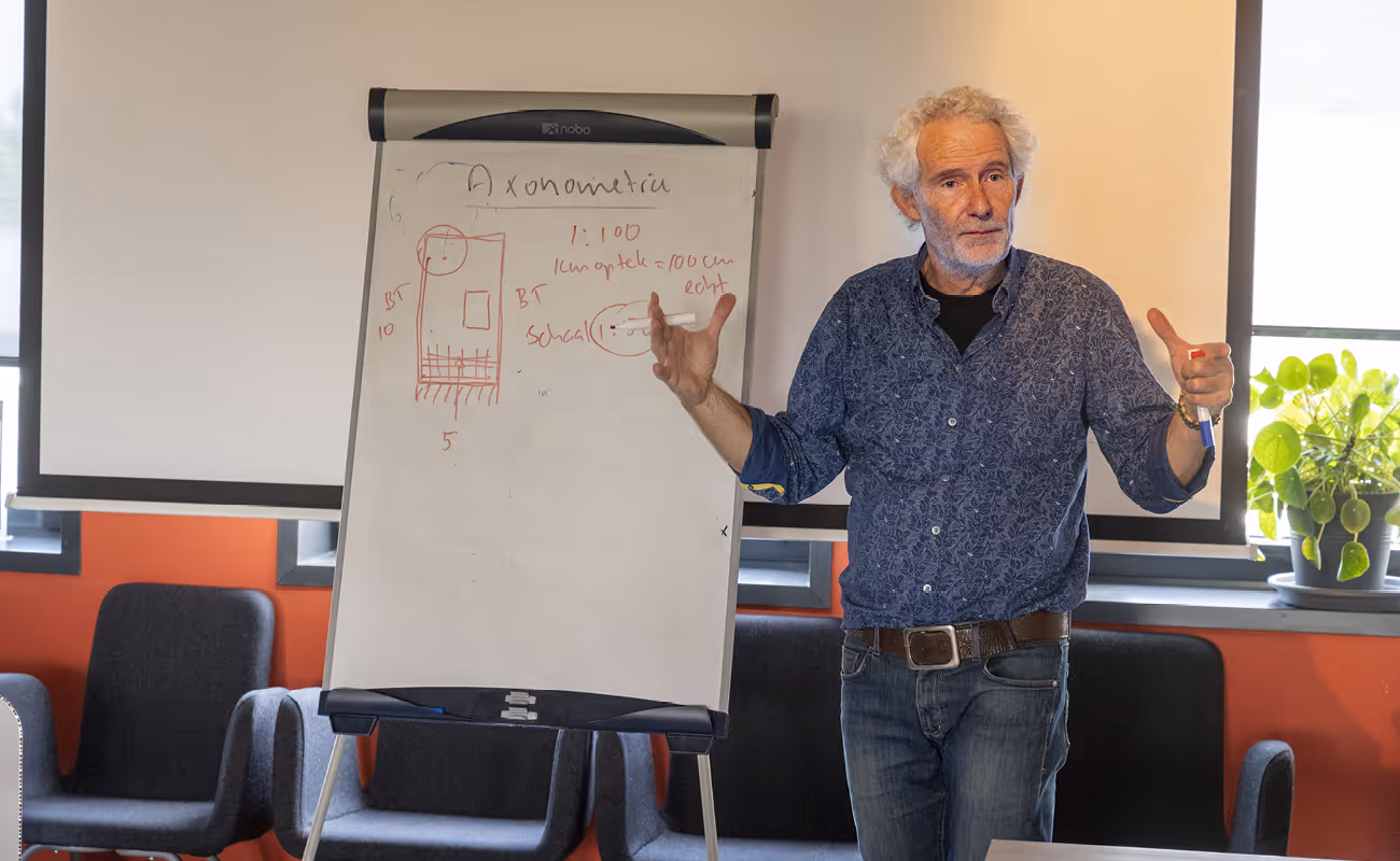 Older man with gray curly hair explaining a diagram on a whiteboard in a meeting room with chairs and a potted plant nearby.