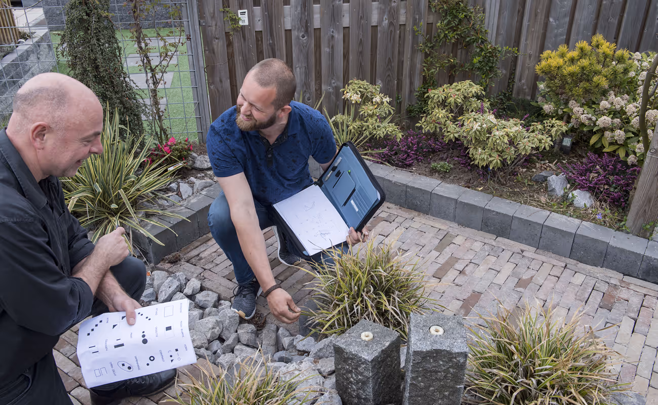 Two men examining landscaping plans and arranging rocks in a garden patio area with plants and a wooden fence.