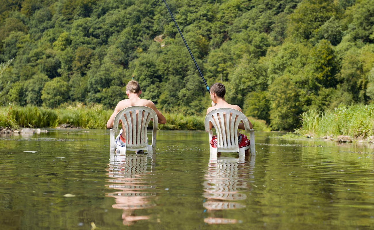 Two boys sitting on plastic chairs fishing in a shallow river surrounded by green trees.