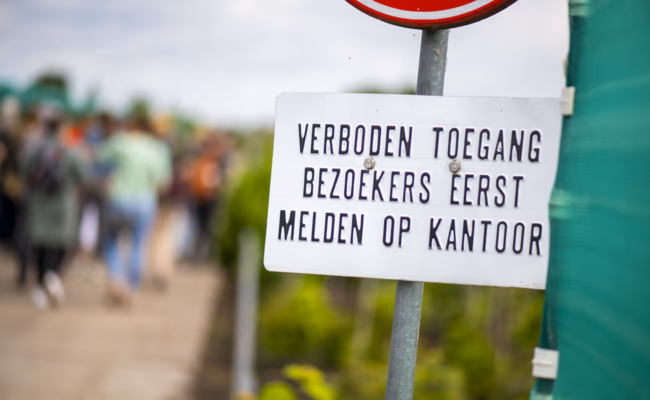 White sign on a metal post reads 'Verboden Toegang Bezoekers Eerst Melden Op Kantoor' with blurred people walking on a path in the background.