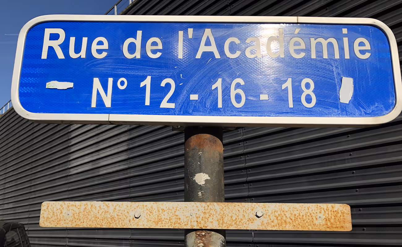 Blue street sign reading 'Rue de l'Académie N° 12 - 16 - 18' mounted on a rusted pole against a black corrugated metal background.