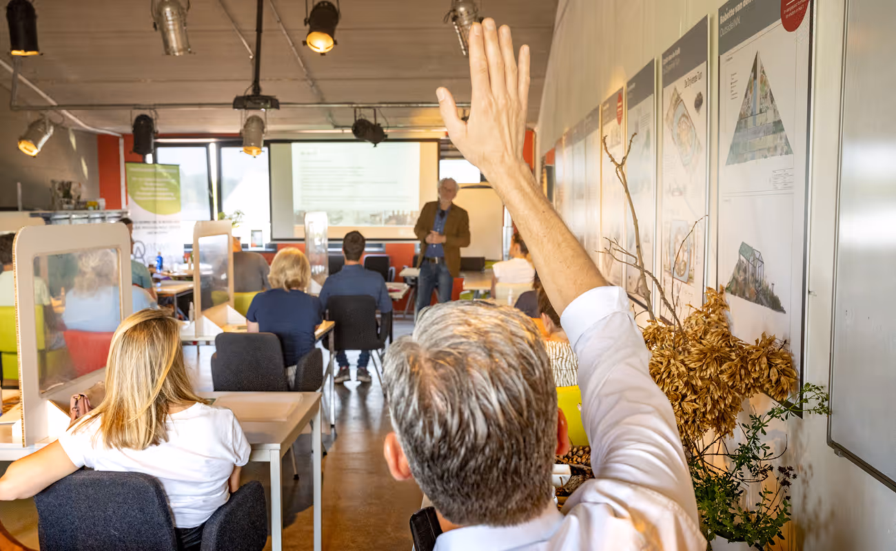 Man raising his hand in a classroom with students facing a lecturer and a presentation screen.