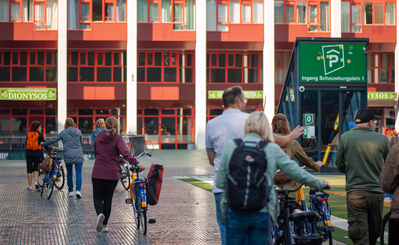 People walking and pushing bicycles on a paved area in front of a red and white building with a green parking entrance sign.