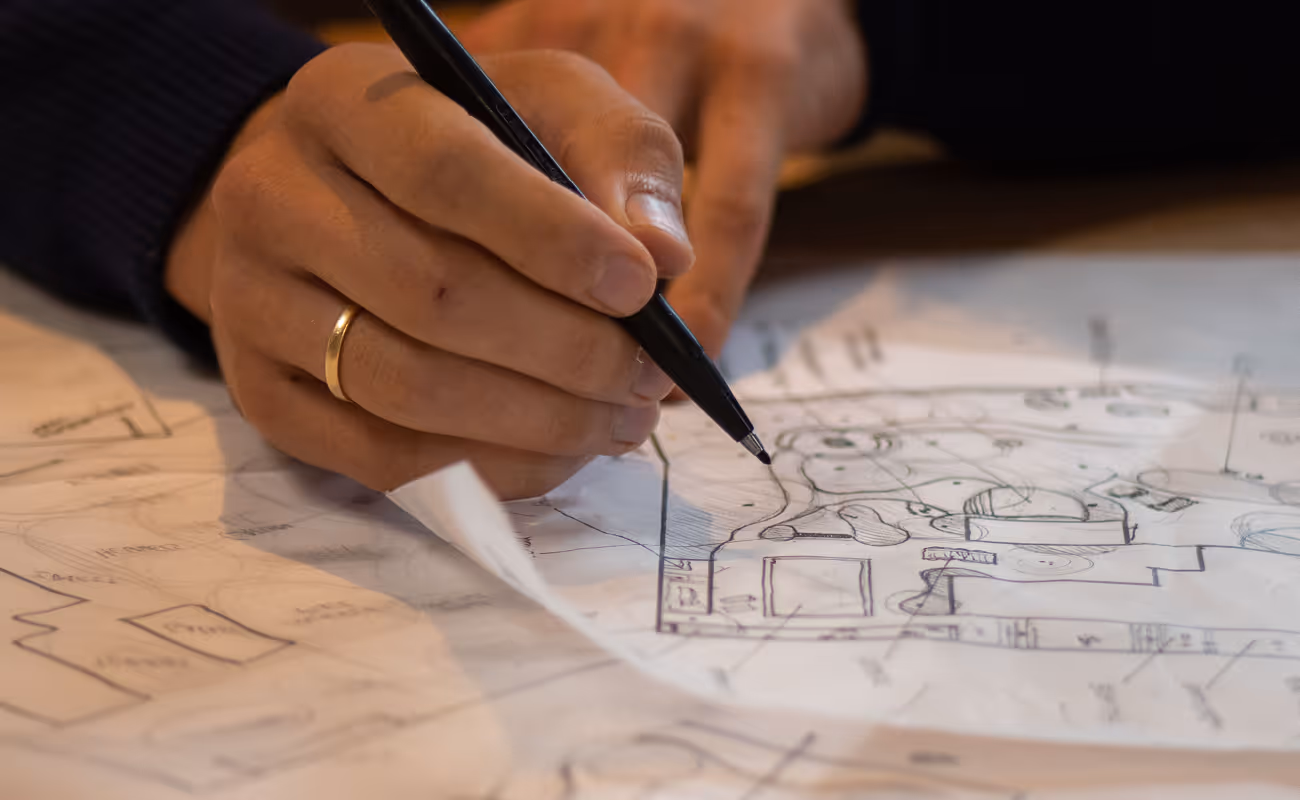 Hand wearing a gold ring holding a pen and sketching on architectural blueprints with detailed floor plans and markings.