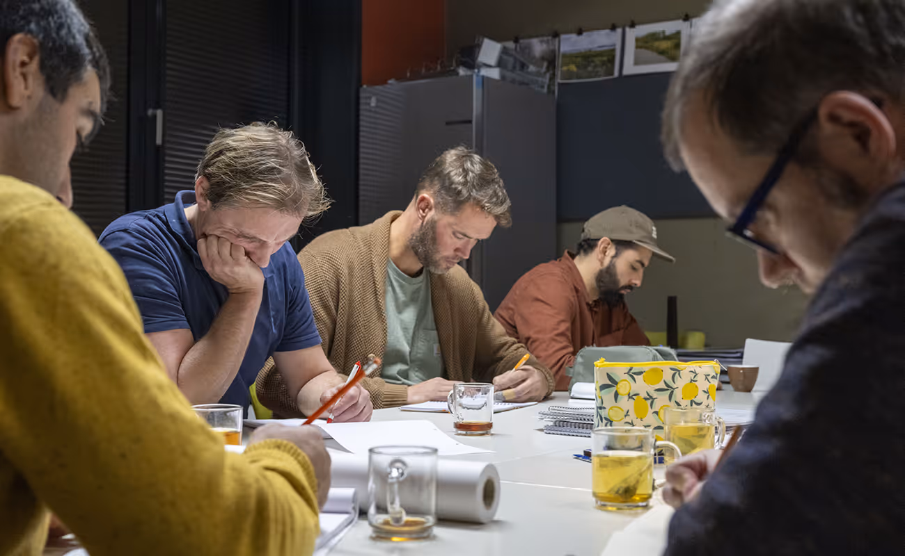 Five men seated around a table concentrating on writing or drawing on papers in a dimly lit room.