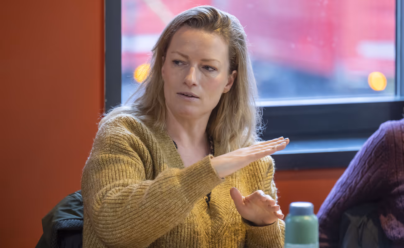 Woman in a mustard sweater gesturing with her hands while speaking indoors near a window.