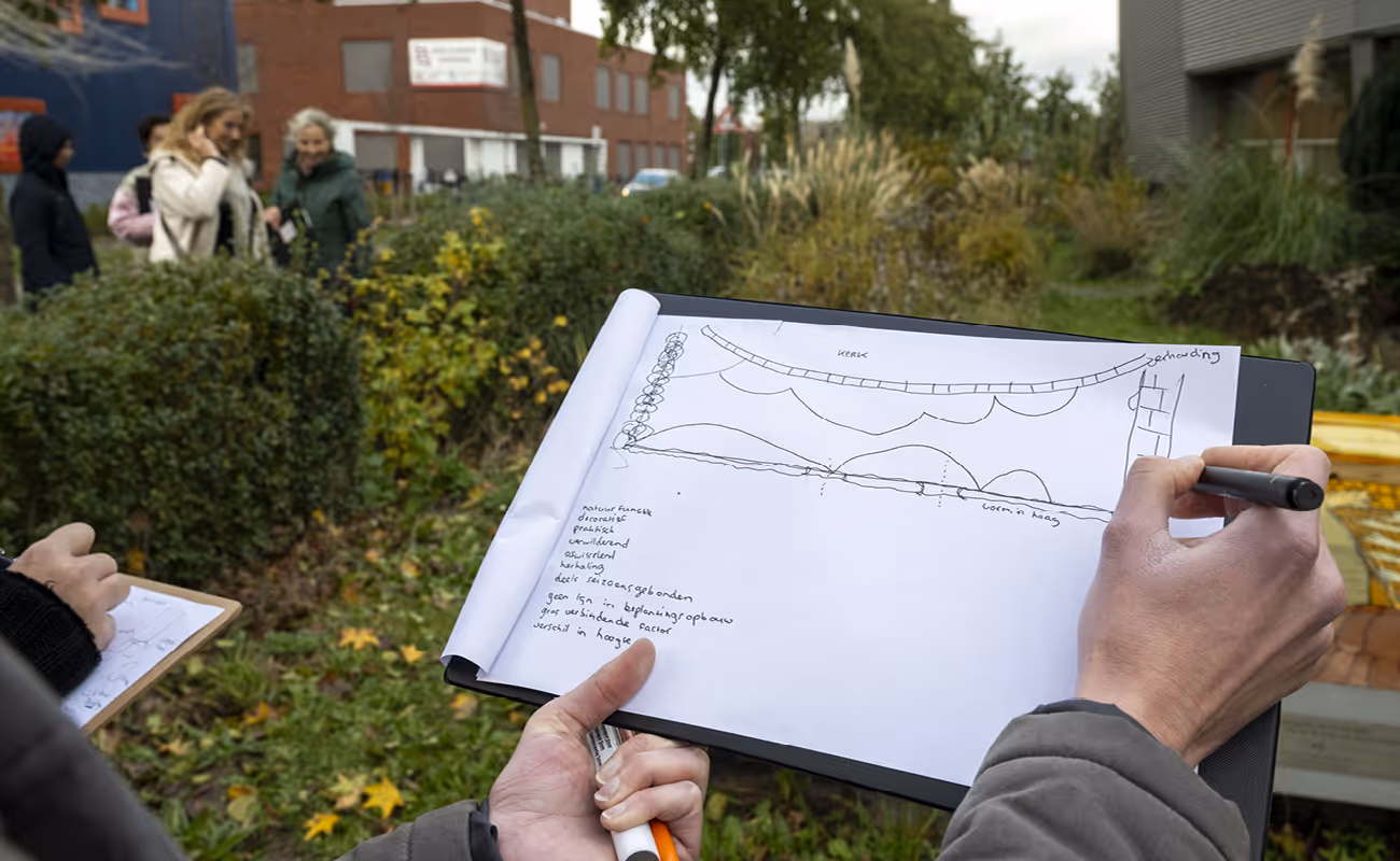 Person holding a clipboard sketching a landscape or garden design with plants, lines, and handwritten notes outdoors.