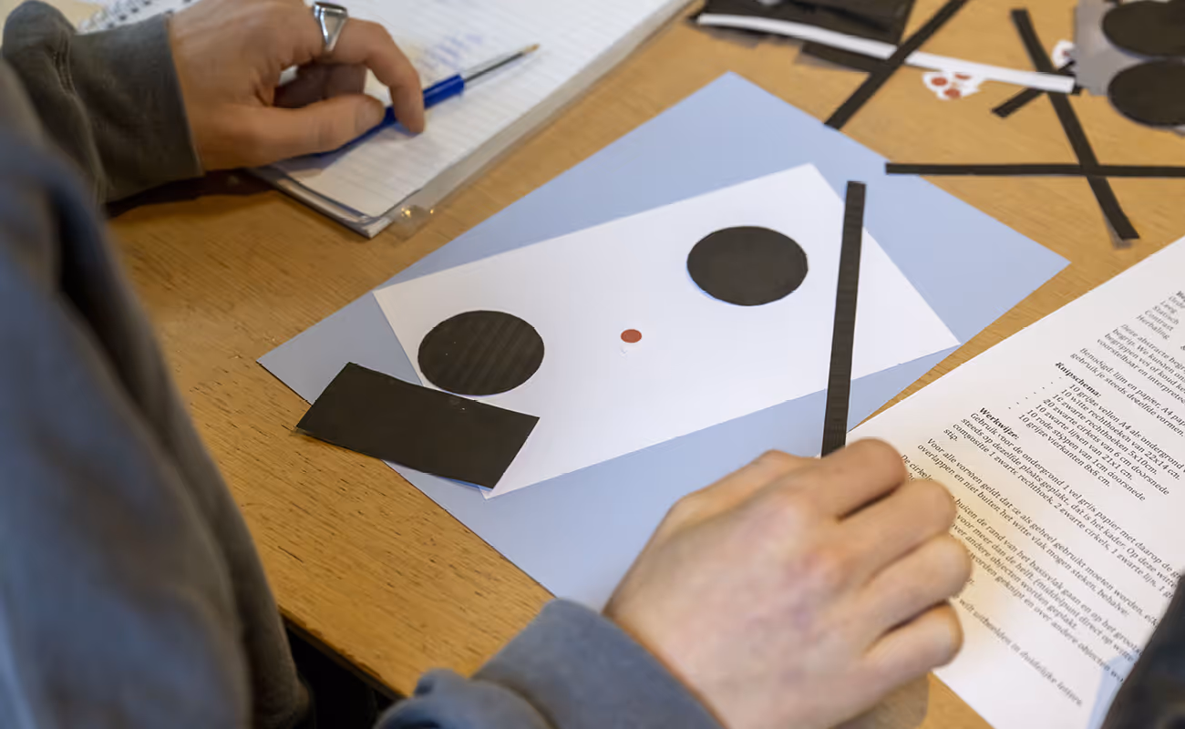 Person assembling black paper shapes on white and blue paper on a wooden table with a pen and notepad nearby.