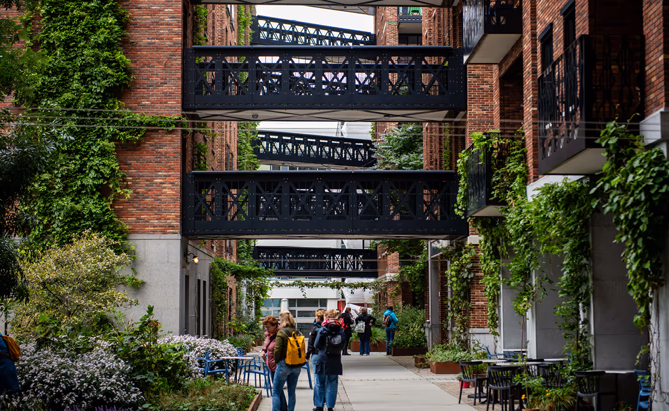 Urban walkway with black iron pedestrian bridges connecting brick buildings covered in green vines and people walking alongside outdoor seating and plants.
