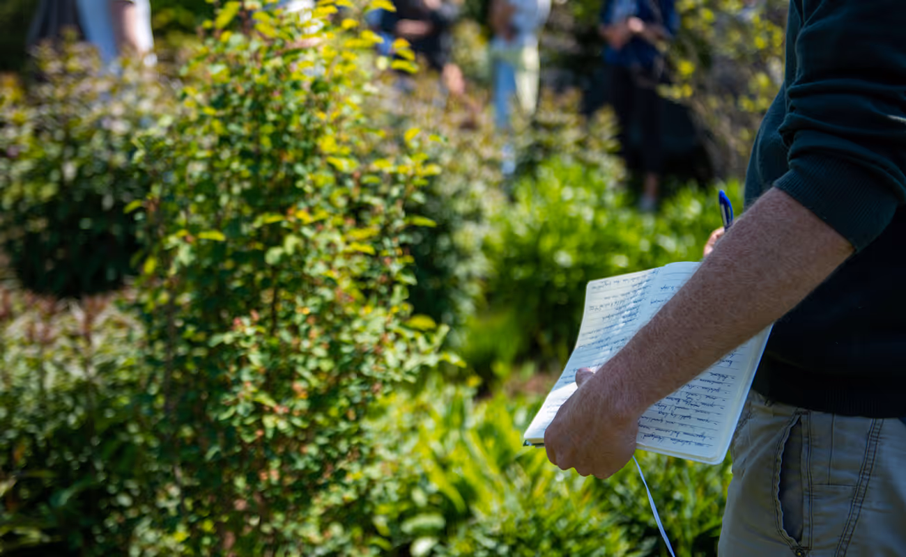 Person holding a notebook with handwritten notes and a pen in an outdoor garden setting.