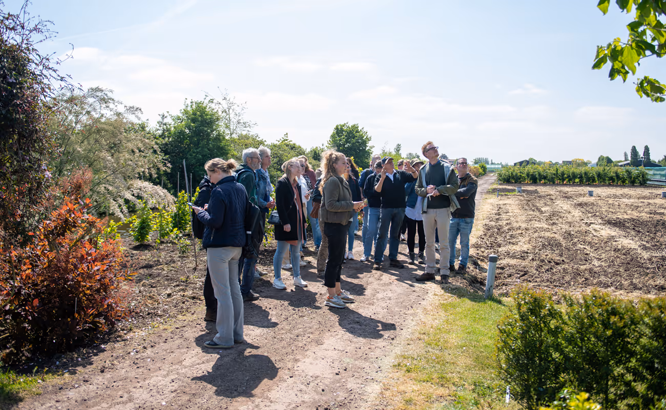 Group of people standing on a dirt path in a garden or farm area with trees and tilled soil under a clear sky.