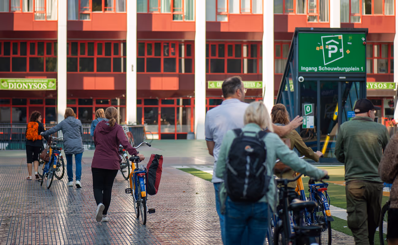 People walking and pushing bicycles near a parking entrance with a green sign reading 'Ingang Schouwburgplein 1' in front of a red and white building.