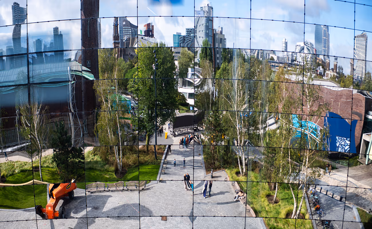 Distorted reflection of an urban park with trees, people, benches, and city buildings on a curved mirrored surface.