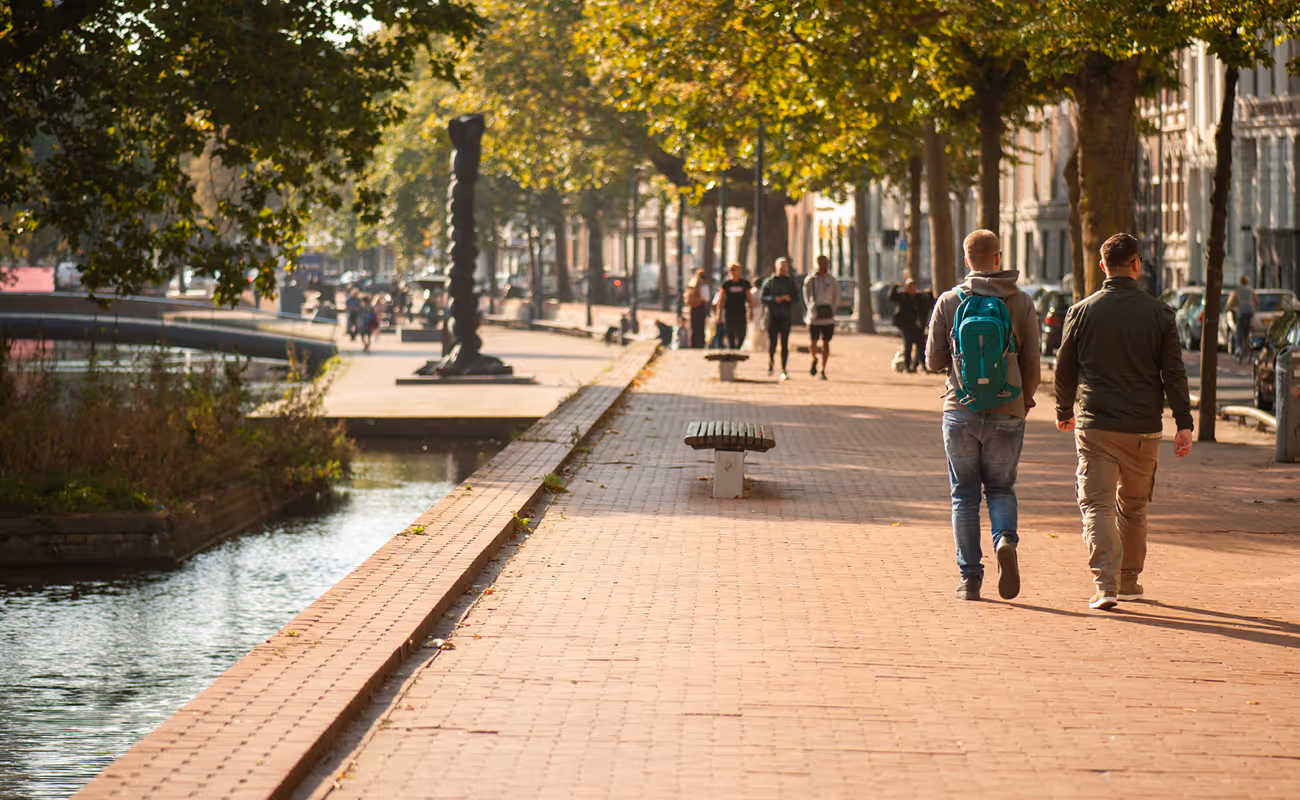 Two men walking along a tree-lined, sunlit canal-side brick pathway with benches and a sculptural column in the background.