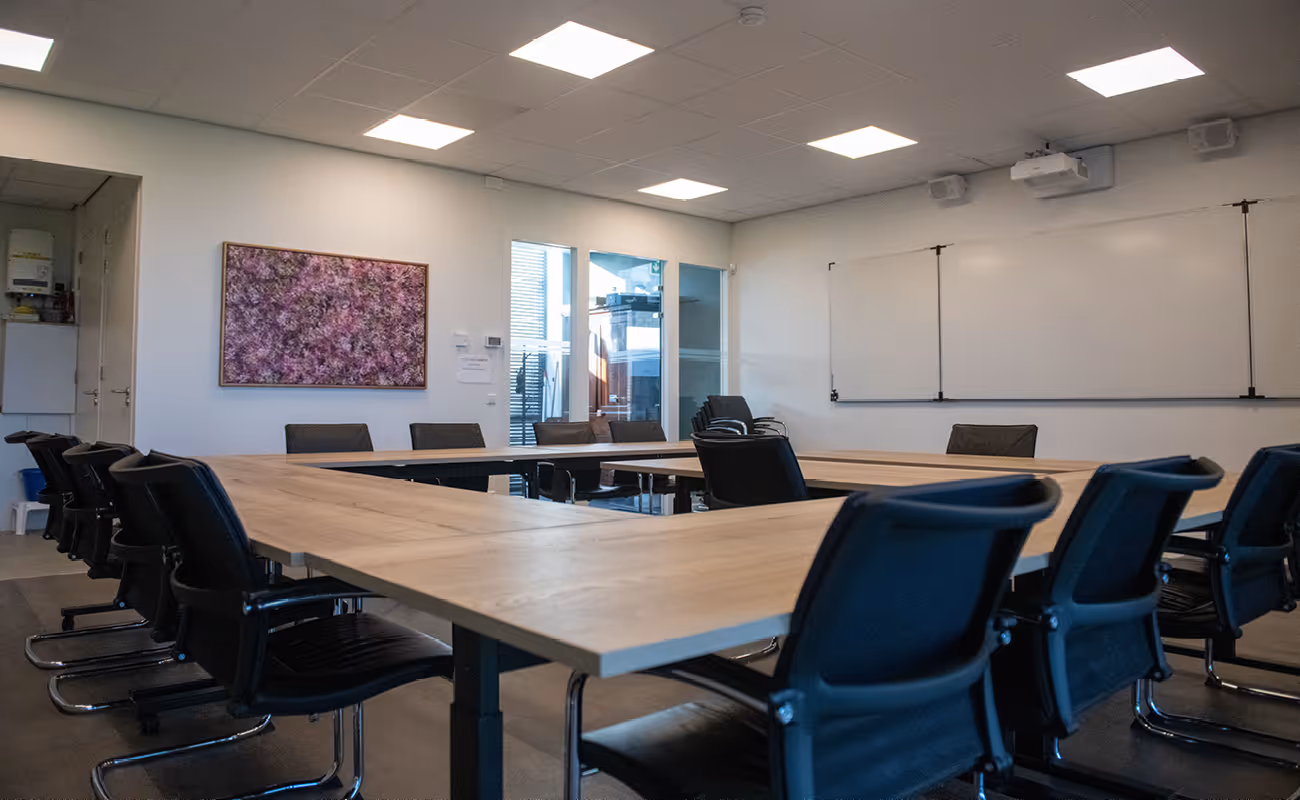 Modern conference room with black office chairs arranged around a large U-shaped wooden table, whiteboard on the wall, and a framed purple floral artwork.