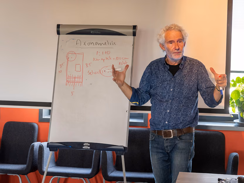 Older man with white hair and beard explaining axonometric drawing on a flip chart in a classroom setting.