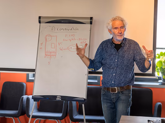 Man with gray hair wearing a blue patterned shirt and jeans gestures while explaining next to a whiteboard with a red diagram and Dutch text in a classroom setting.