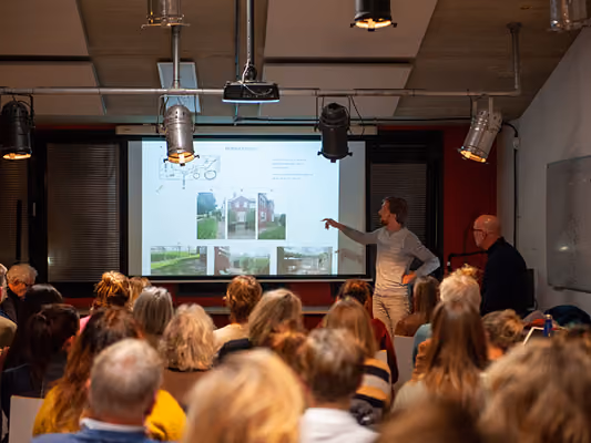 An audience watches a man presenting architectural images and plans projected on a screen in a conference room.
