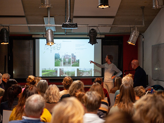 An audience watches a man presenting architectural images and plans projected on a screen in a conference room.