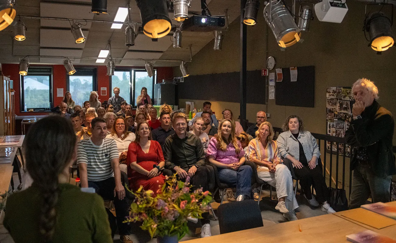Audience attentively listening to a speaker in a dimly lit room with flowers on the table in front.