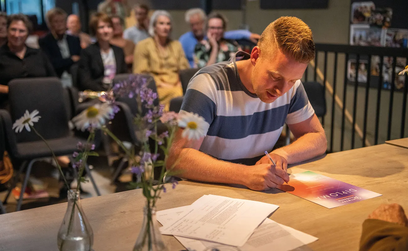 Man signing a colorful diploma at a table with papers and flowers, with an audience watching in the background.